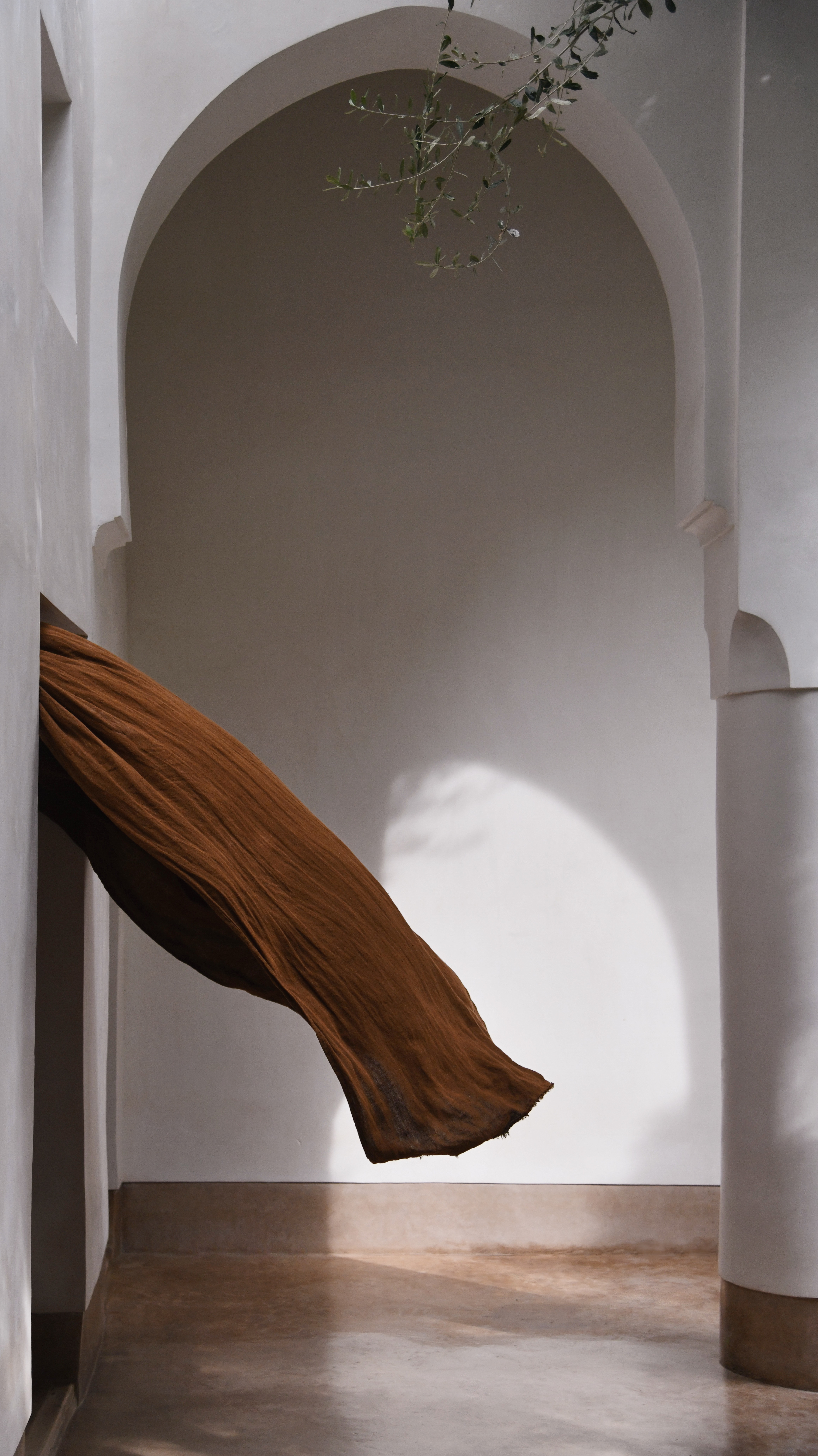 Brown fabric cascades through an arched doorway in an empty white hallway with a tan floor and a leafy branch hanging overhead.