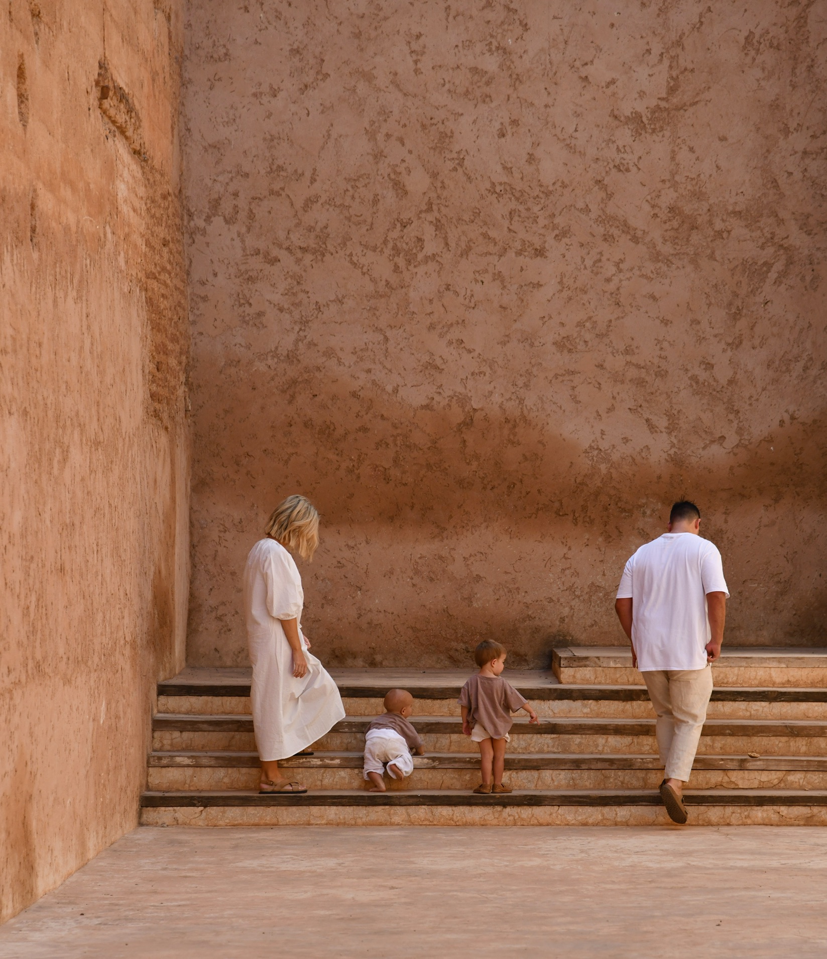 Family of four walks on stairs in a courtyard, with light-tan walls and natural light. Parents and children on vacation, exploration.