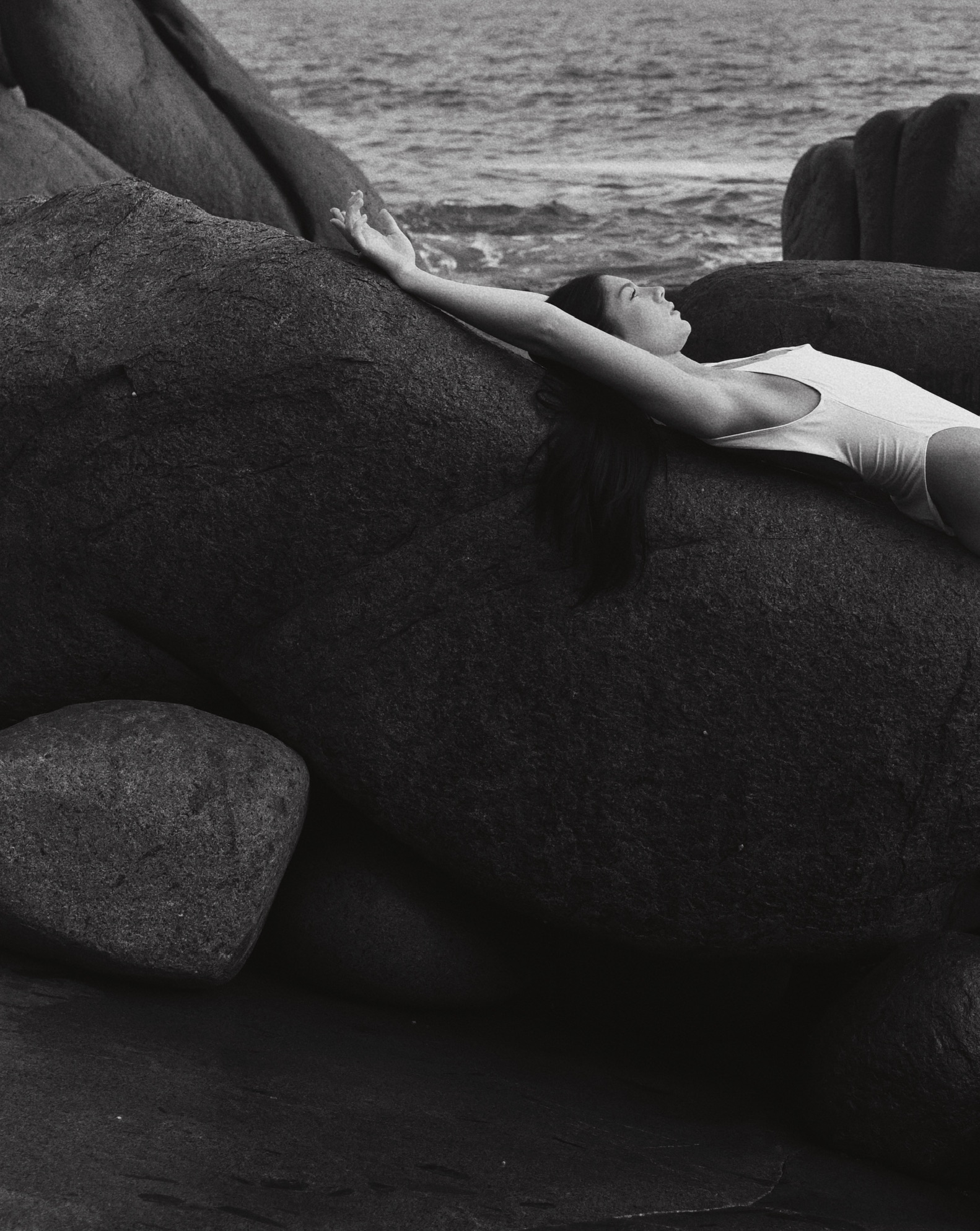 Woman in white swimsuit lying on large rocks by the ocean, arms stretched out overhead, black and white, contemplative, peaceful.
