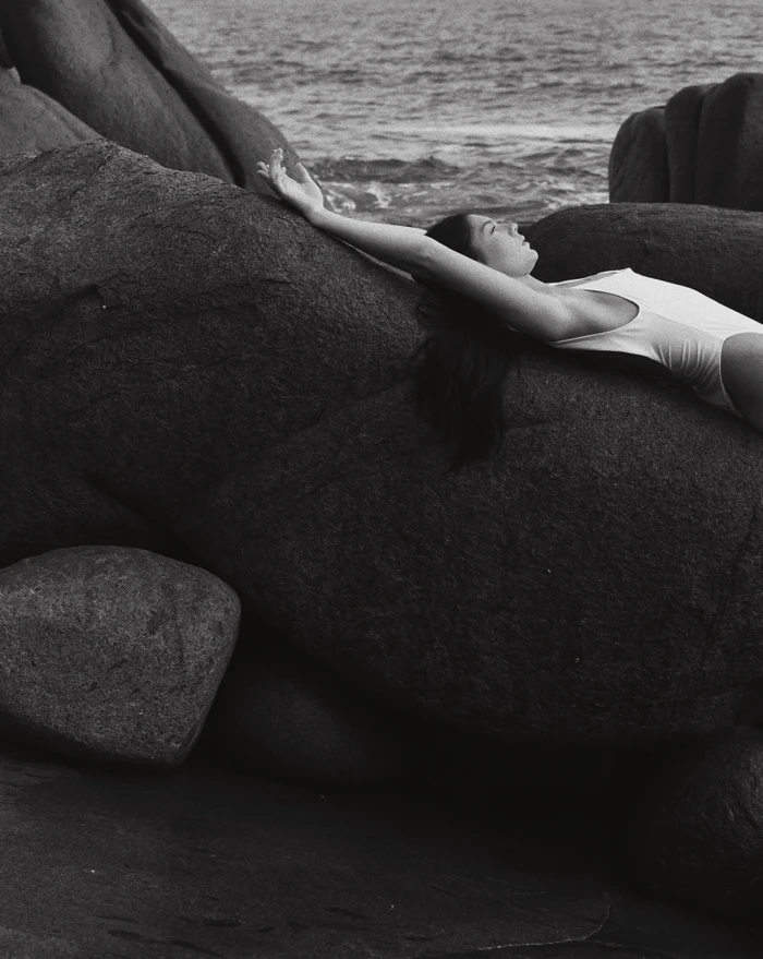 Woman in white swimsuit lying on dark rocky coastal boulders by the ocean photographed by Sarah Jessica Marie Burns.