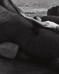Woman in white swimsuit lying on dark rocky coastal boulders by the ocean photographed by Sarah Jessica Marie Burns.