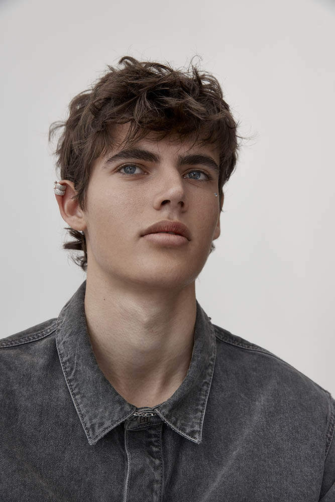Portrait of a young adult male with brown hair, blue eyes, wearing a gray denim shirt against a plain, neutral background.