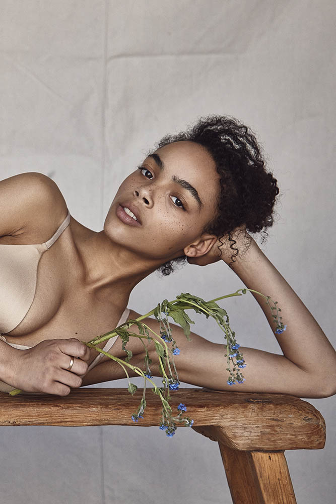 An adult female with curly hair reclines indoors on a wooden surface with a bouquet of blue flowers in a natural-light beauty shot.