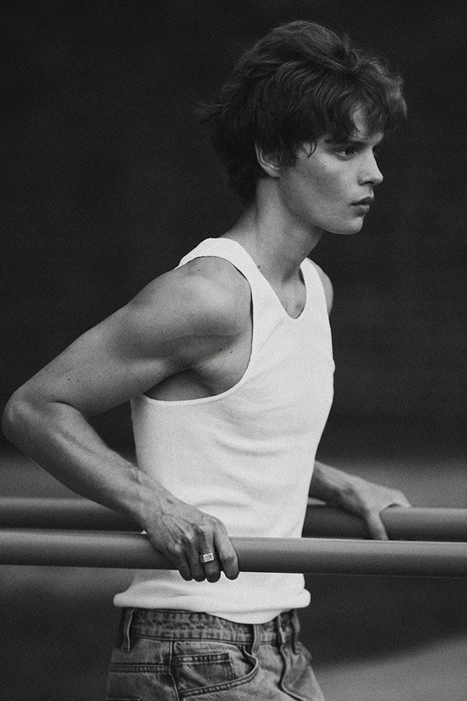 Black and white portrait of a young adult male in a white tank top and blue jeans holding onto a metal bar outside in daylight.