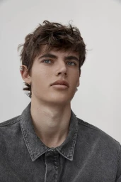 Close-up portrait of a young man with dark wavy hair wearing a grey denim shirt against a neutral background.