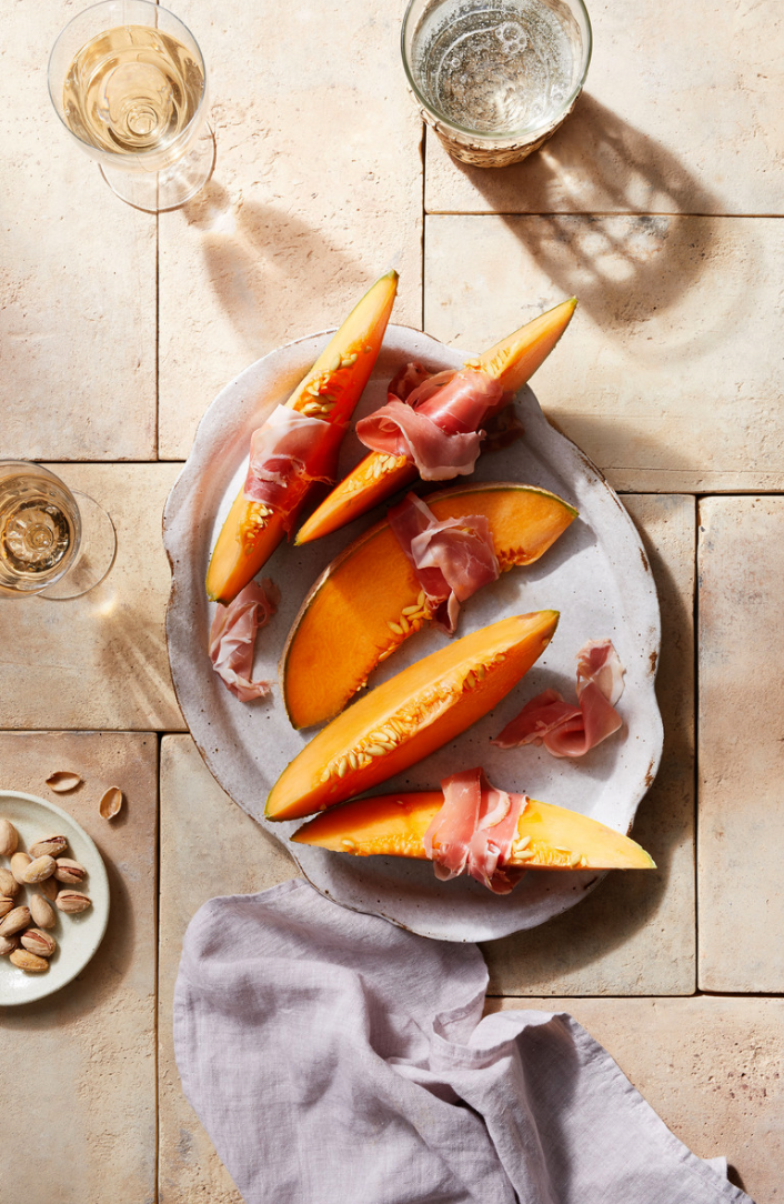 Overhead shot of cantaloupe wedges wrapped in prosciutto on a plate with drinks and pistachios against a tile background.