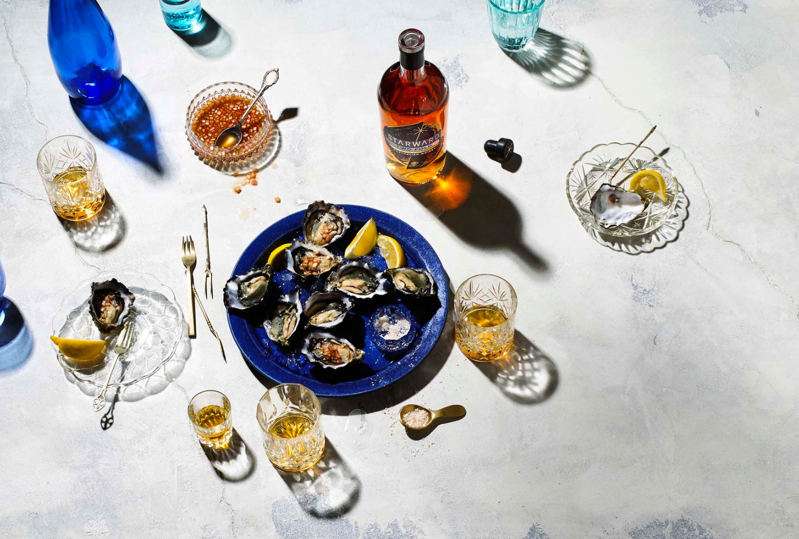Overhead shot of oysters on blue plate with lemon wedges, whiskey glasses, bottle, and sauce on marble background with shadows.
