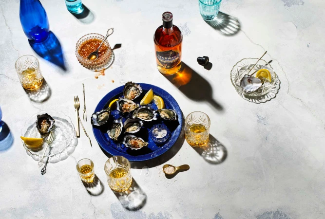 Overhead shot of oysters on blue plate with lemon wedges, whiskey glasses, bottle, and sauce on marble background with shadows.