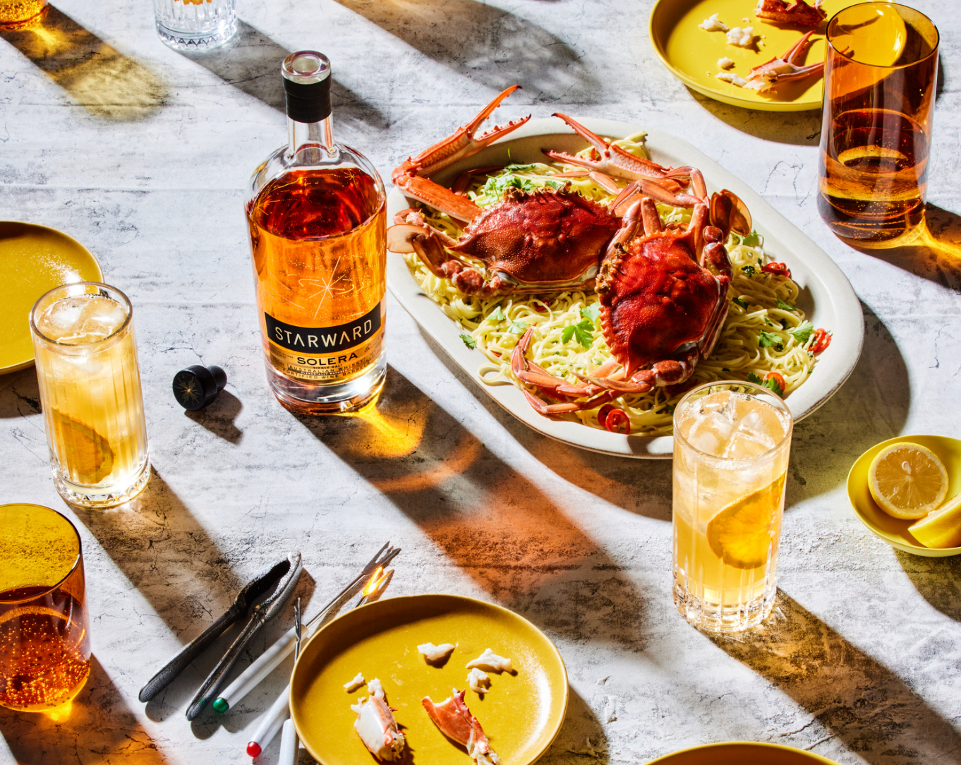 Overhead shot of a food and beverage still life with pasta, crab, cocktails, and a bottle of Starward Solera whiskey.