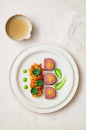 Overhead food styling shot of sliced meat roulade, shredded root vegetables, green dollops, and a small ceramic bowl of broth.