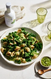 Overhead food styling shot of pasta with kale pesto in a white bowl with a bottle of oil and glassware on a marble table.