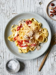 Plate of yellow pasta with red tomatoes and grated cheese on a distressed white wooden table with a gold fork