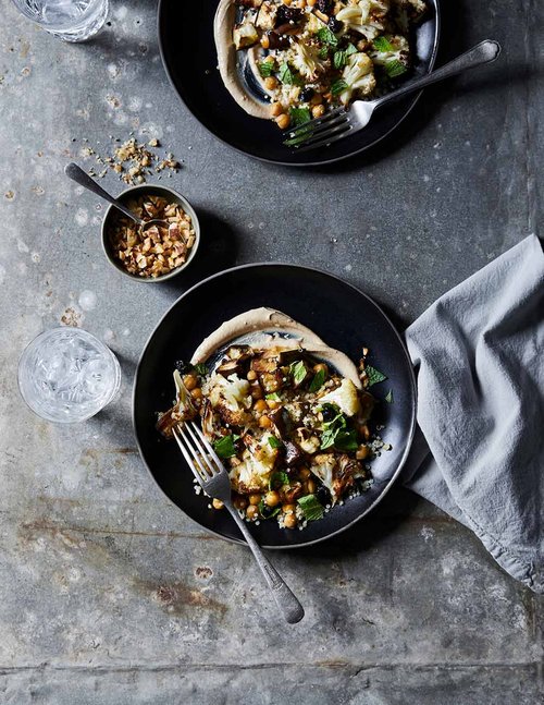 Overhead shot of two black plates of roasted cauliflower with tahini, chickpeas, and herbs, plus a small bowl of nuts on a gray surface.