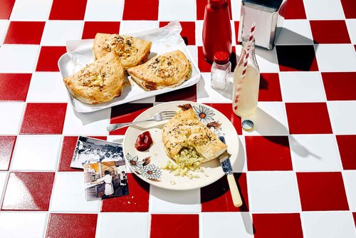Overhead shot showcases half-eaten savory pies on a floral plate, accompanied by side dishes and condiments, set against a red and white checkered surface.