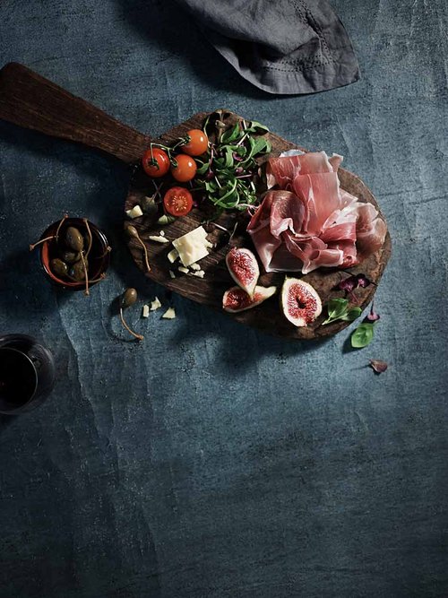 Overhead shot of a rustic wooden cutting board with cured ham, figs, cheese, tomatoes, olives and herbs on a textured blue surface.