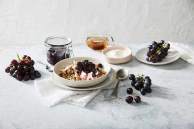 Bowl of granola with yogurt and berries on a marble surface with loose grapes, glass jar, and tea cup styled by Peta Gray