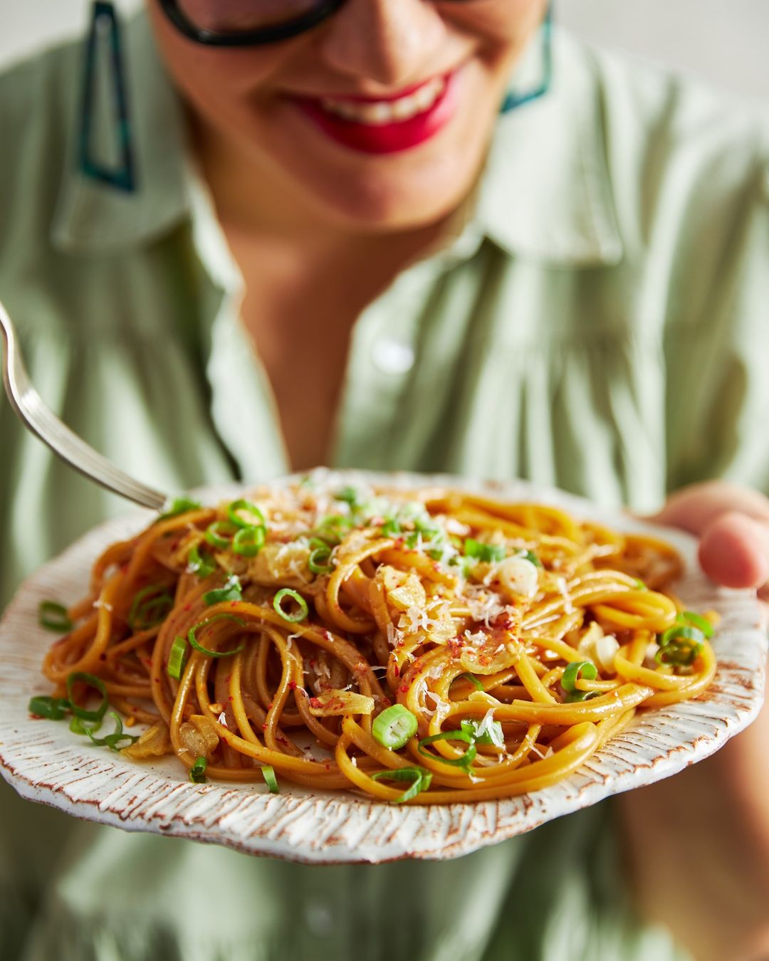 Close-up of a person holding a plate of spaghetti with scallions and parmesan cheese, ready to eat, in a bright indoor setting.