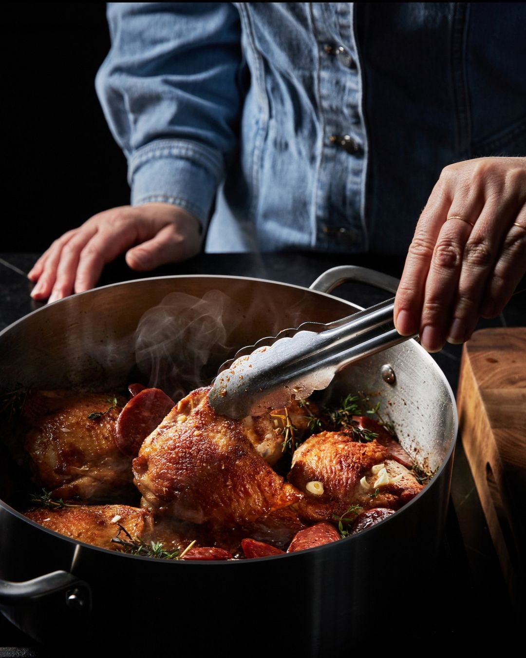 Adult cooking chicken thighs and sausage in a pot, indoors, using metal tongs, wearing a denim shirt, with steam rising.
