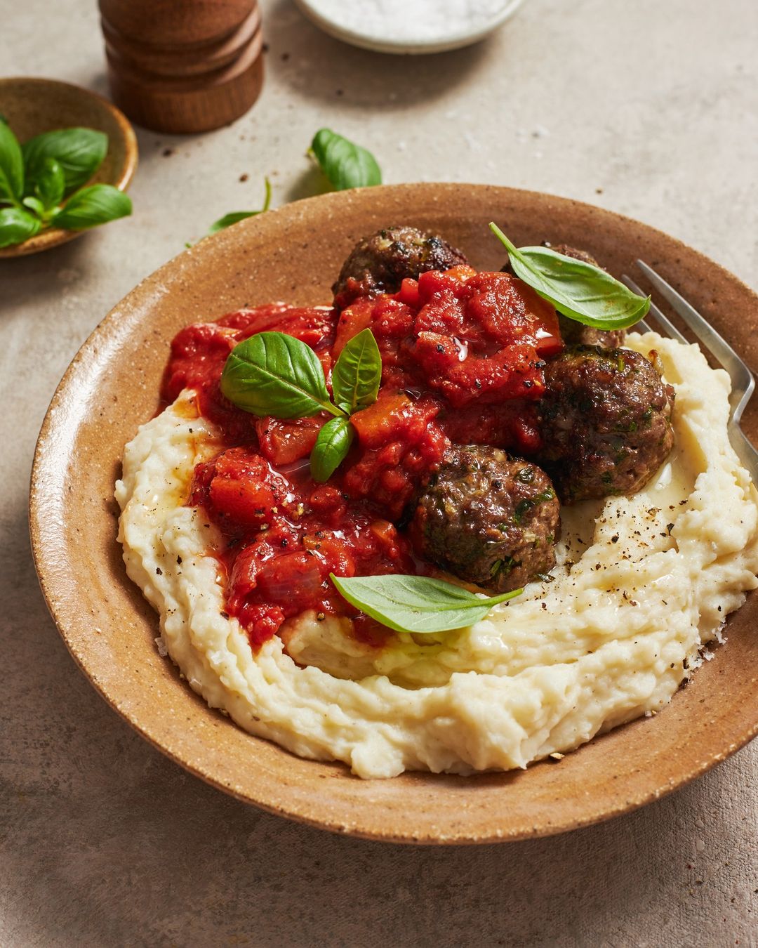 Overhead shot of meatballs with tomato sauce and mashed potatoes on a brown plate, garnished with basil leaves on a beige surface.