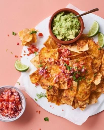 Top-down view of loaded nachos with melted cheese, fresh salsa, guacamole, and lime wedges on white paper