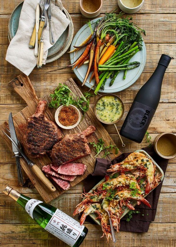Overhead shot of two grilled steaks, grilled shrimp, sake bottles, and various side dishes on a rustic wooden table, arranged for a meal.