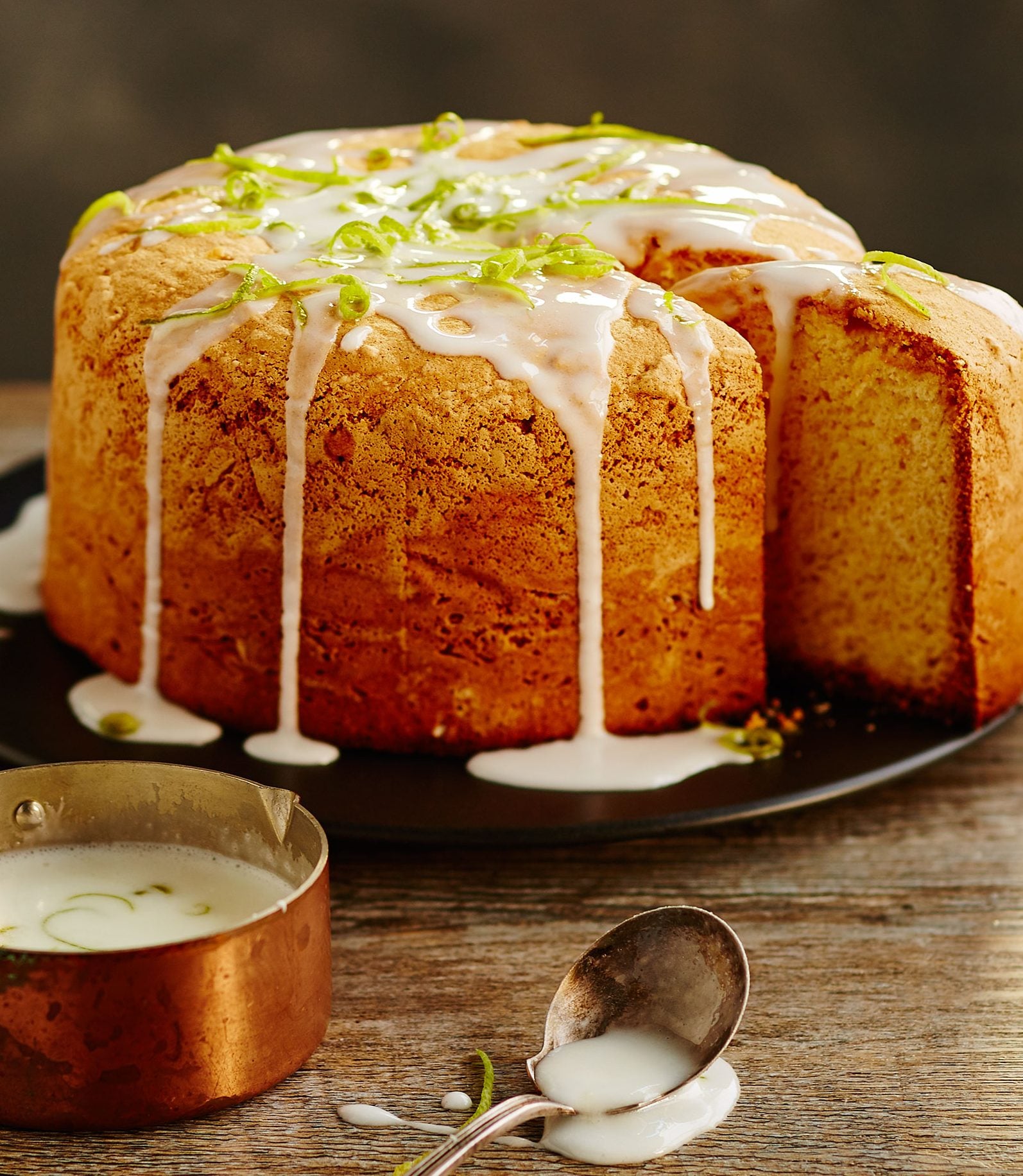 Close-up studio shot of a ring cake on a black plate with white icing and lime zest, accompanied by a spoon and a copper pot filled with white sauce.