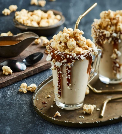 Two caramel popcorn milkshakes with straws on a bronze serving tray with popcorn bowl and wooden board with a caramel sauce container.