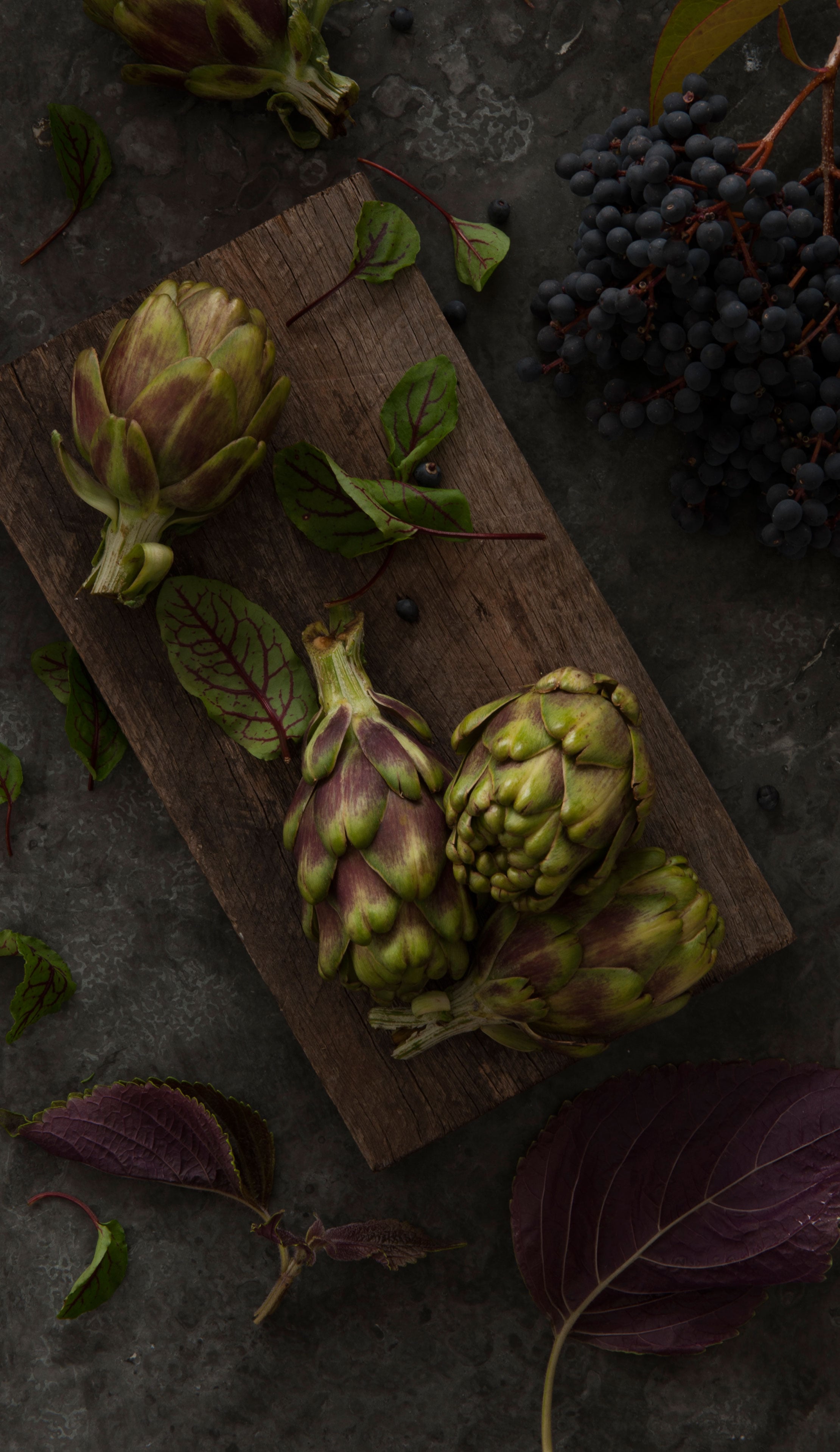 Overhead shot of artichokes on wooden board, arranged with dark berries and leaves on a dark grey surface. Food and beverage photo.