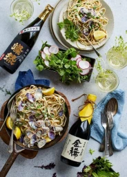 Top-down view of two pasta dishes with clams, fresh salads, and two bottles of sake on a light grey textured surface.