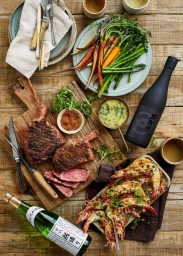 Top-down food styling featuring grilled steak on a cutting board, roasted vegetables, and wine bottles on a wooden table.