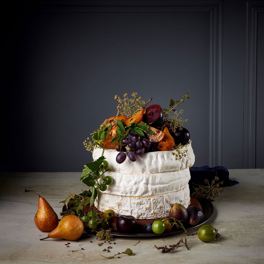 Stacked brie cheese wheel cake topped with fresh fruit including pears, grapes, plums and figs against a grey wall.
