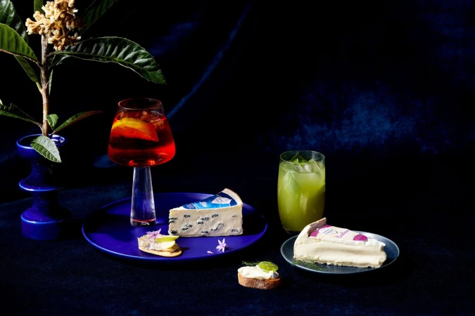 Still life photo of two glasses of drinks, two cheese slices, and canapés on a dark blue surface and background, with a floral branch.