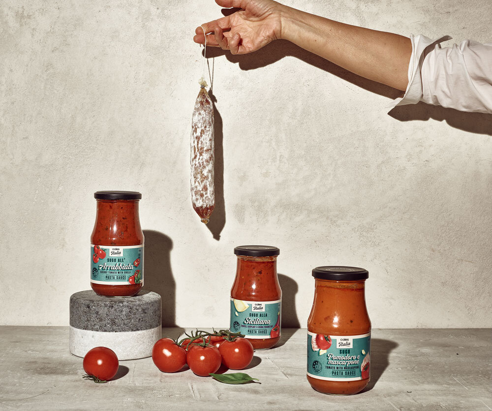 Three jars of tomato sauce sit next to tomatoes, with a hand holding salami against a light gray wall in a well-lit indoor setting.