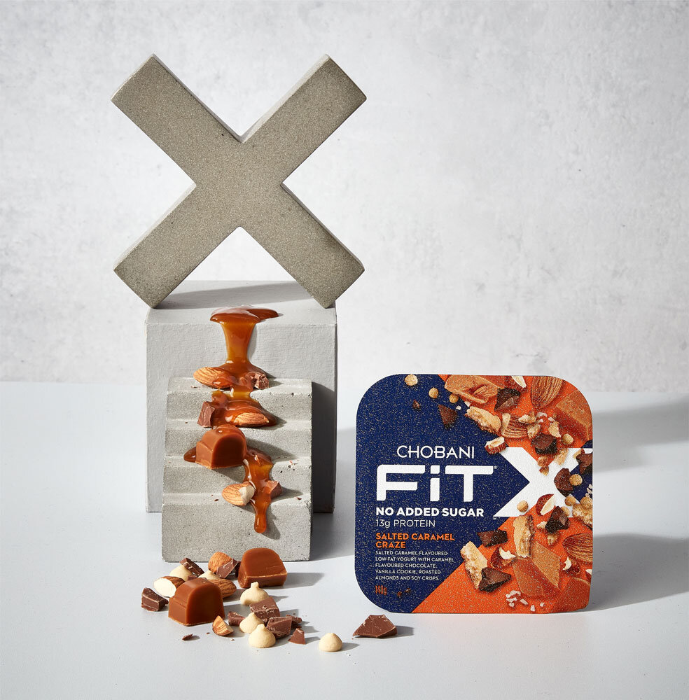 Chobani Fit X Salted Caramel Craze container surrounded by caramel candies and almonds on a white surface with a geometric backdrop.