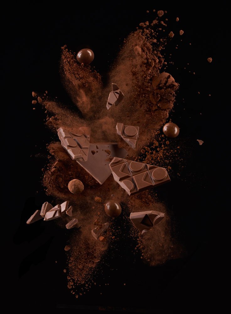 Flying milk chocolate chunks and spheres explode with cocoa powder against a black backdrop, creating a visually appealing food still life.