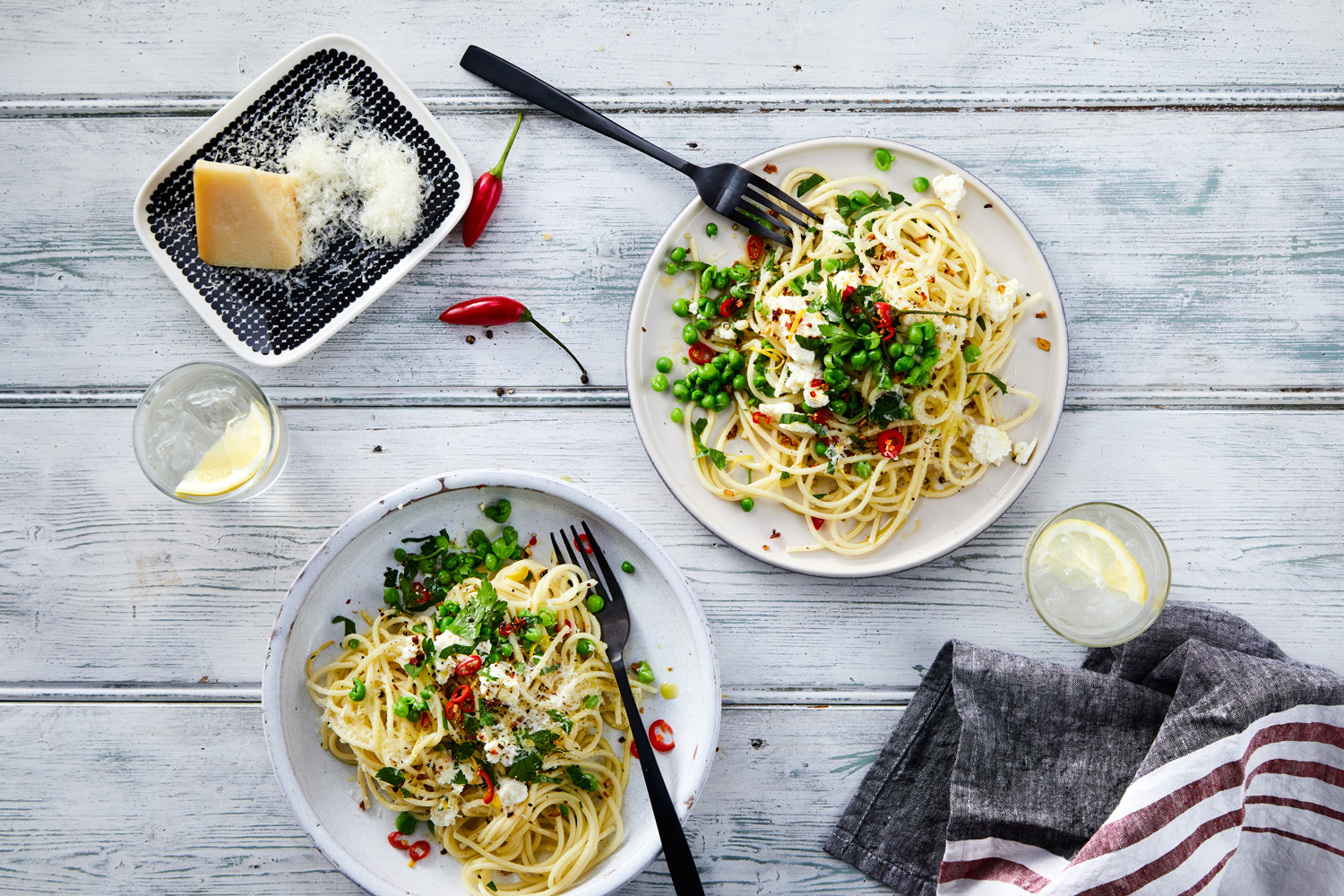 Overhead shot of two plates of spaghetti with peas, cheese, herbs and peppers, served with water and a side of cheese.