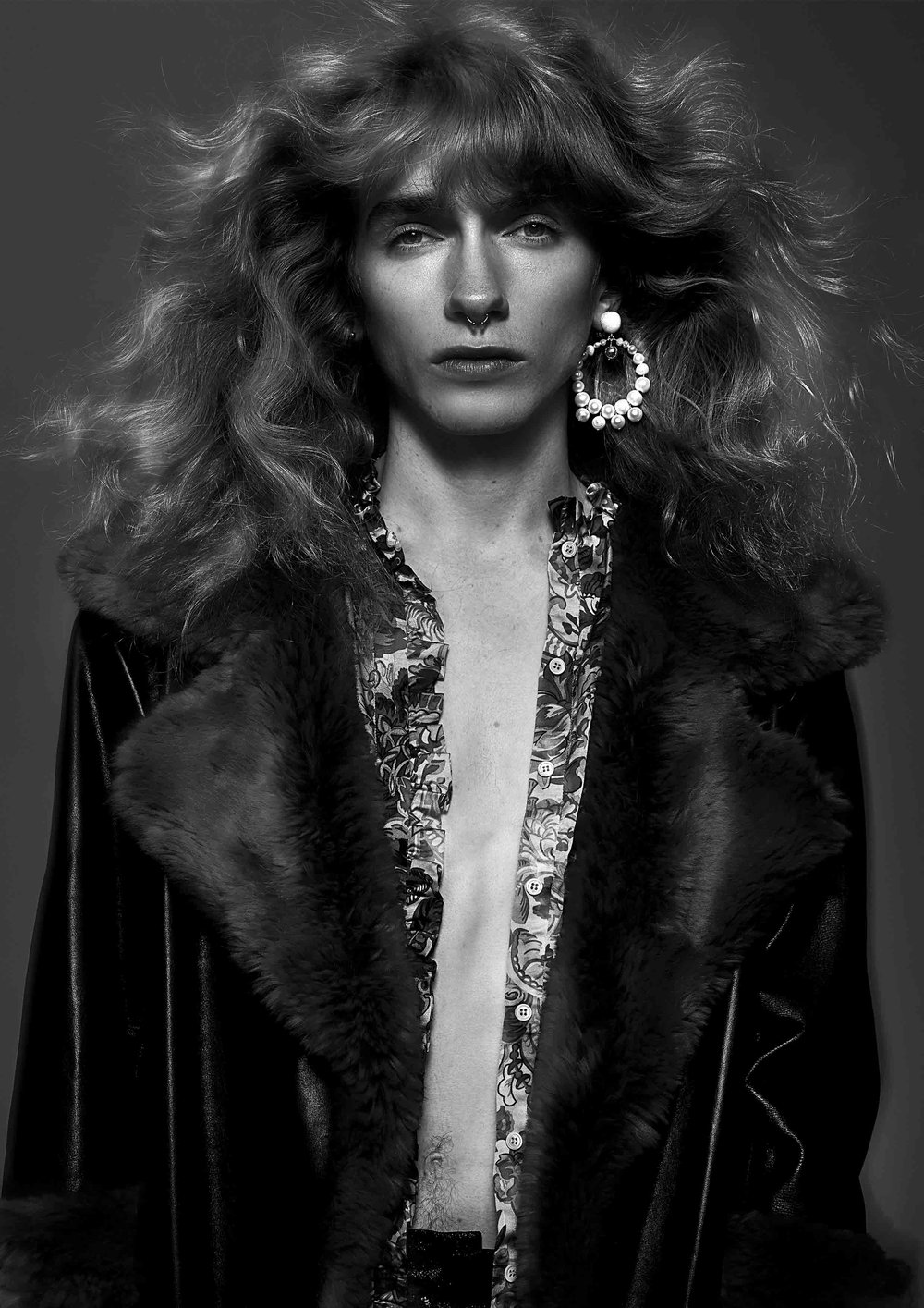 Portrait of a person in a black and white studio shot, wearing a fur-lined coat and floral shirt, with long wavy hair and pearl earrings.