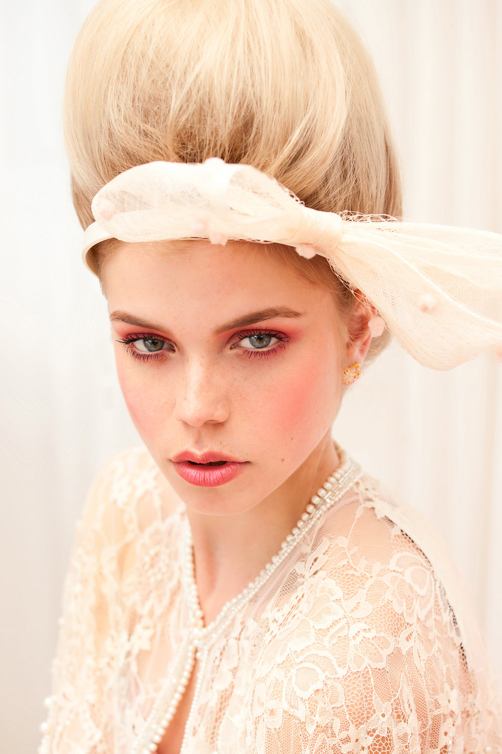 Close-up portrait of a blond woman with elaborate makeup and a lace top, set against a bright white background in a beauty shot.