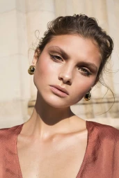 Close-up portrait of a woman with braided updo hair, warm makeup, and gold earrings wearing a terracotta top against an outdoor building background