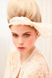 Close-up portrait of a woman with high blonde bouffant hair and a lace headband, wearing a sheer white lace dress.