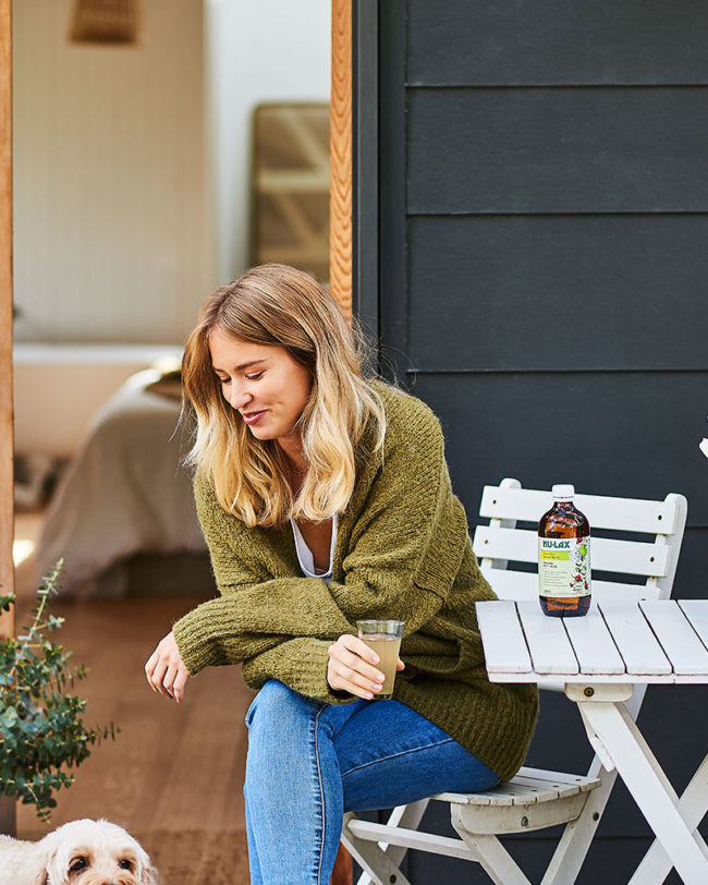 A blonde woman in a green sweater sits at a white table outdoors, drinking from a glass with a dog nearby and a bottle on the table.