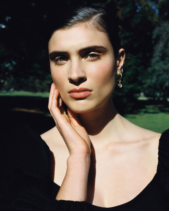 Portrait of a young woman with dark hair and fair skin, posing outdoors in soft light. She wears a dark top with a natural makeup look.