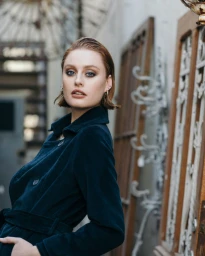 Portrait of a woman with wet-look reddish-brown hair wearing a dark blue coat outdoors with wooden panels.
