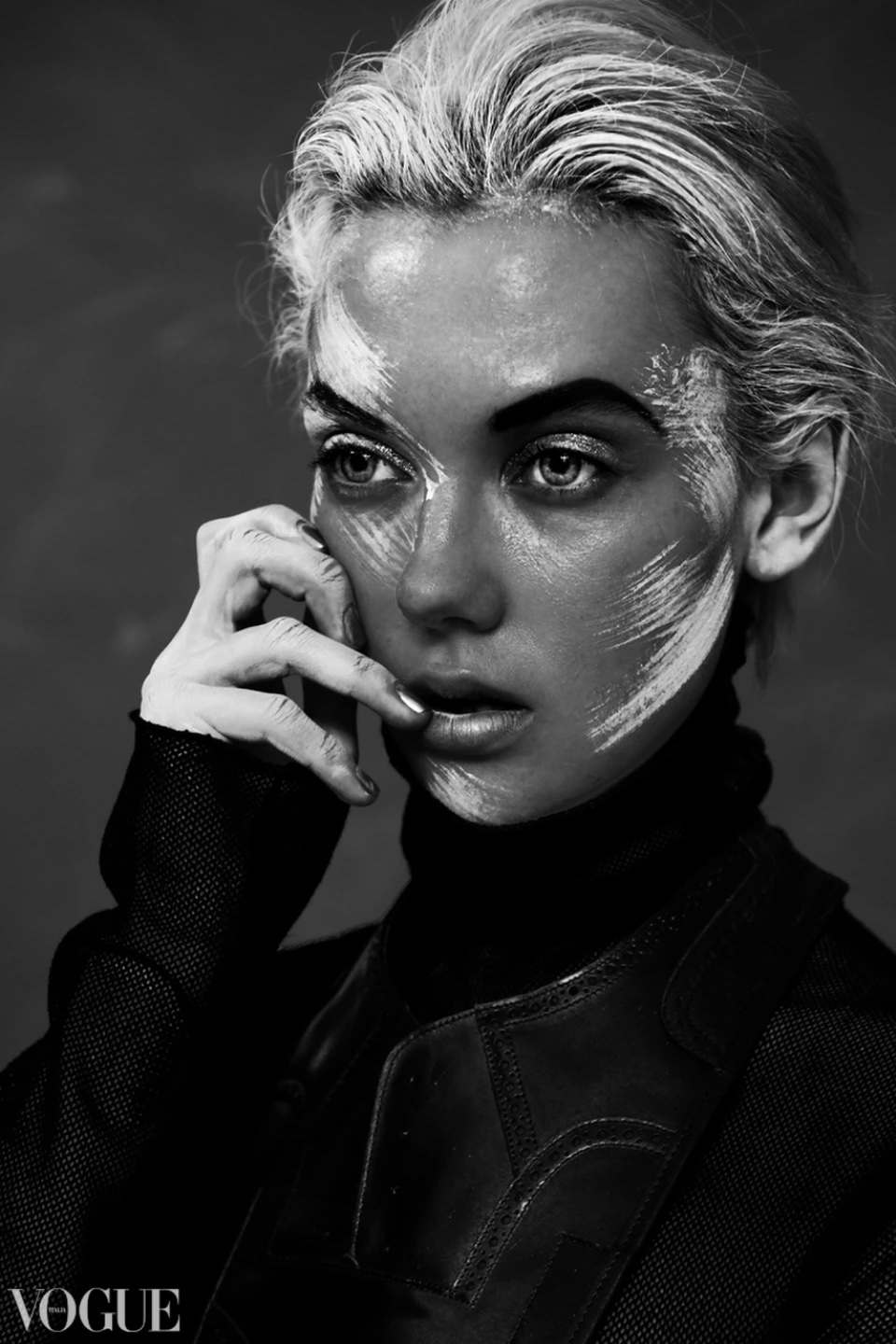 Monochrome portrait of a young adult woman with white painted lines on her face, wearing a turtleneck and blazer, Vogue logo visible.