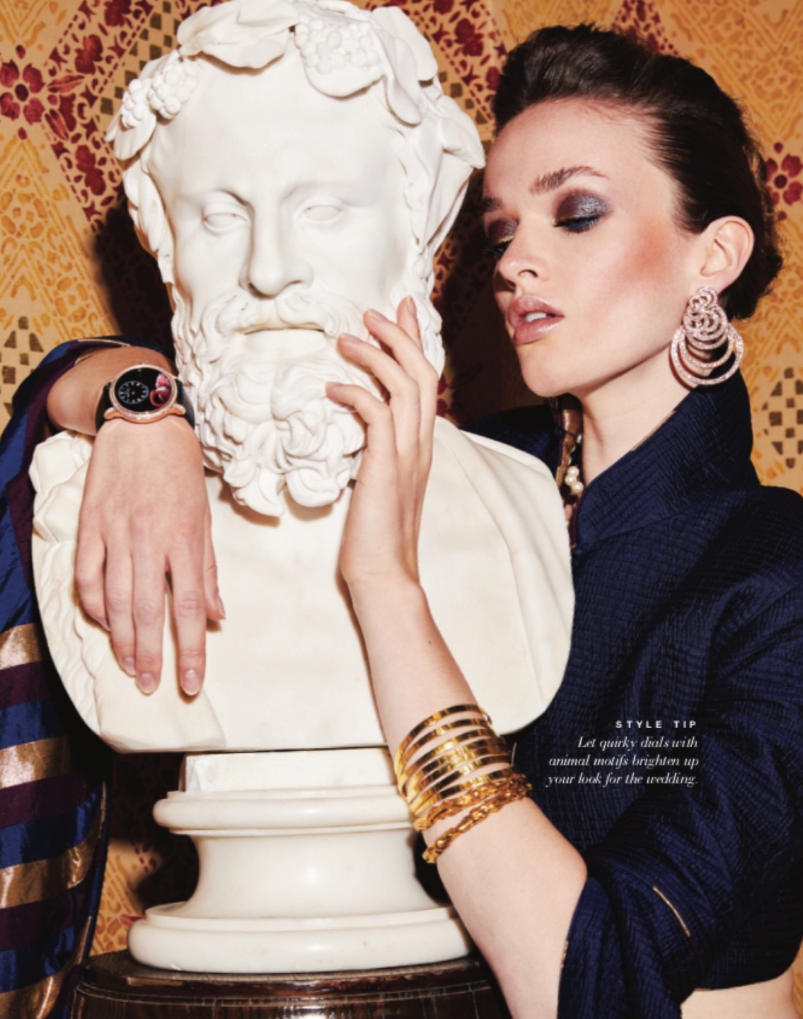 Elegant woman in a navy blazer poses with a classical bust sculpture and gold jewellery, against a patterned damask background.