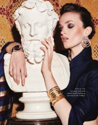 Woman wearing gold hoop earrings and layered bracelets posing beside a white classical marble bust statue.