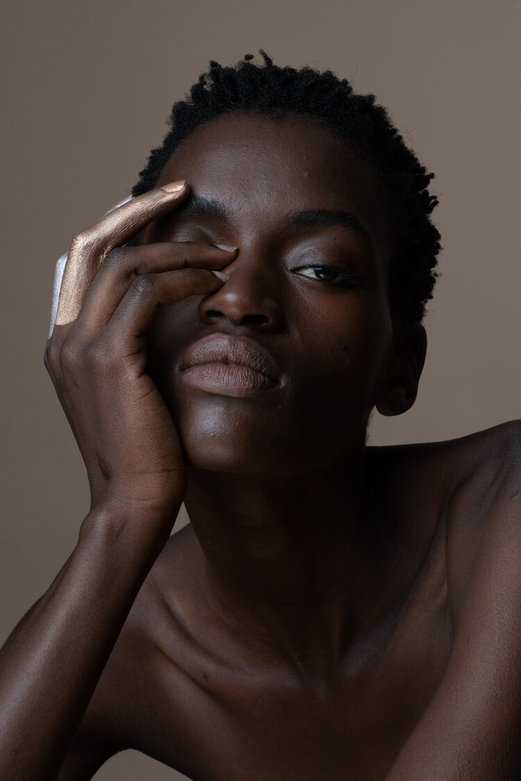 Close-up portrait of a dark-skinned woman with short hair, one hand painted gold partially covering her eye, against a tan background.
