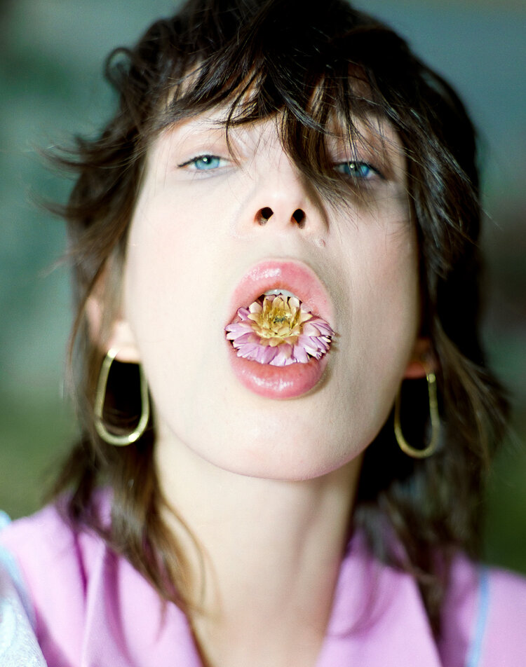 Close-up of a young woman with dark hair holding a pink flower in her mouth, wearing gold hoop earrings and a lavender shirt.