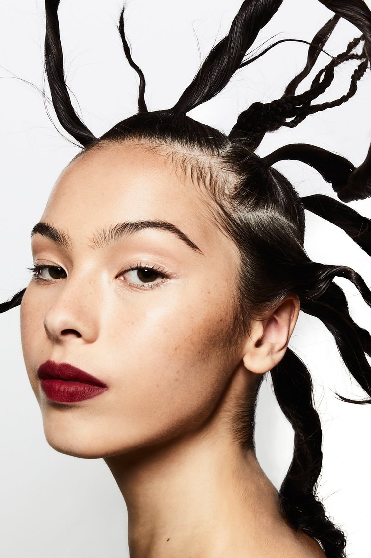 Close-up portrait of a young woman with dark burgundy lipstick and a unique hairstyle of twisted strands against a white background.