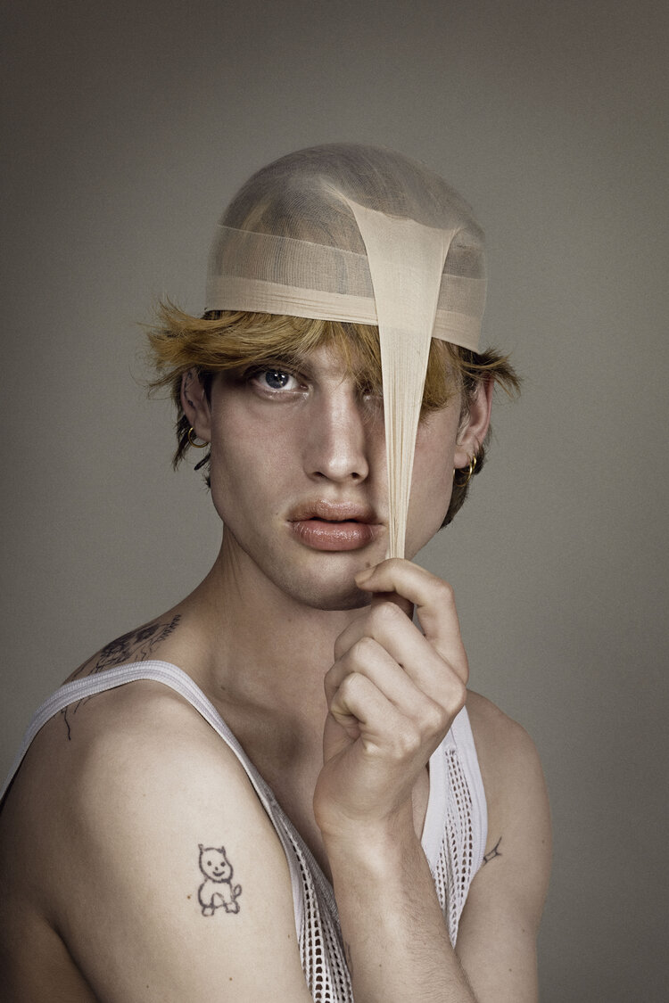 Portrait of a young adult male with blonde hair, wearing a white tank top and nude-colored stocking cap indoors in front of a neutral wall.